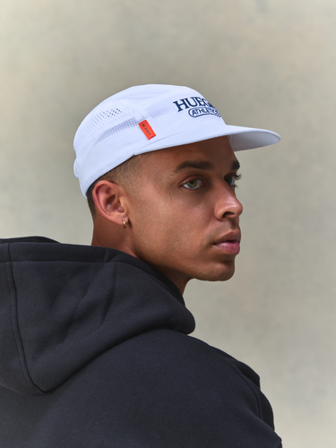 A man in a black hoodie wearing a white Huega running cap, shown in a side profile against a textured beige background.