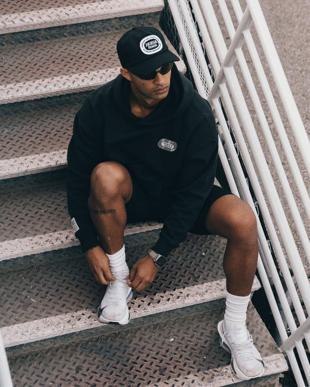 Man sitting on stairs wearing a black hoodie, shorts, and white sneakers.