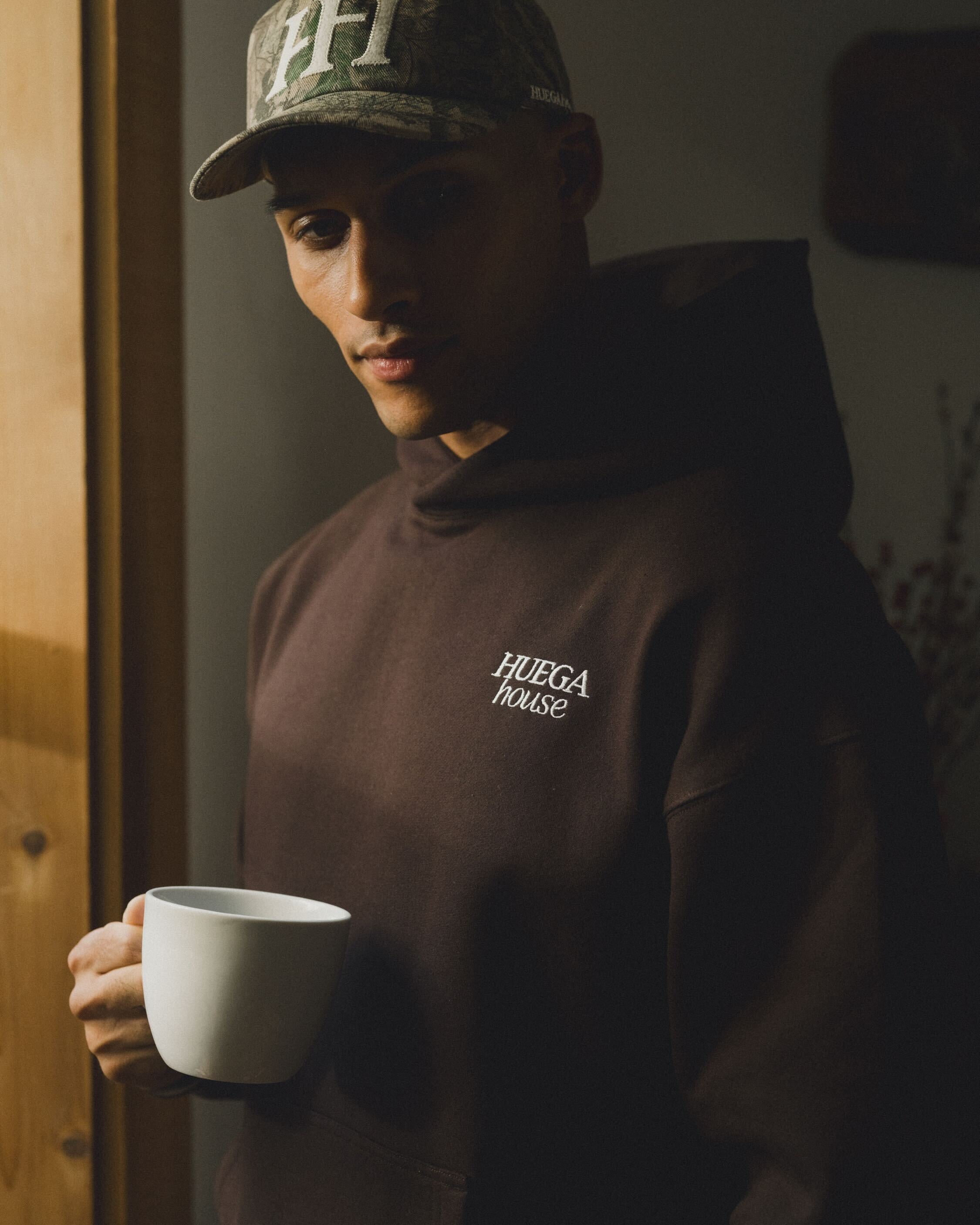 Person wearing a hoodie with 'HUEGA HOUSE' logo holding a white mug indoors.