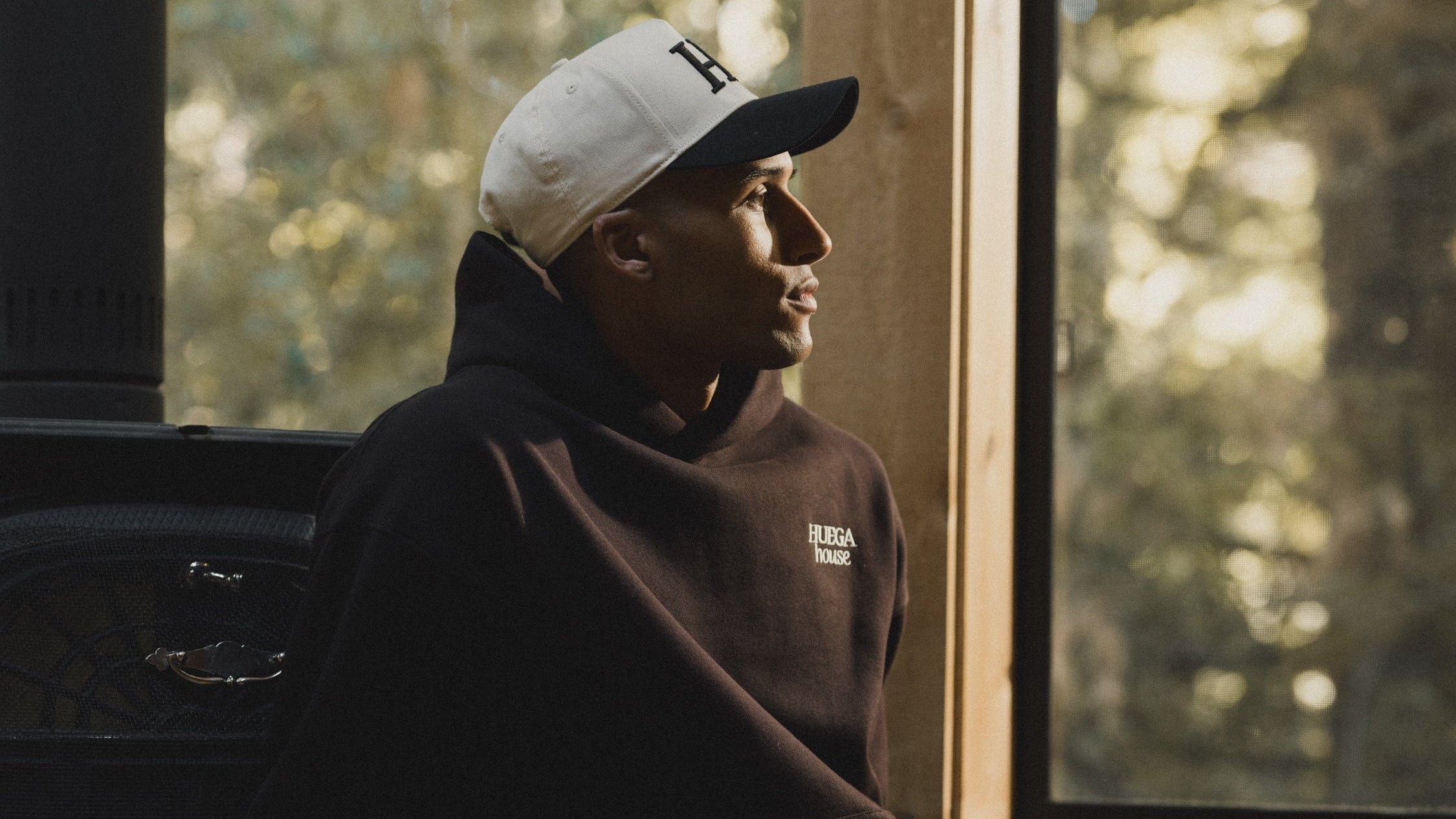 Person wearing a hoodie and cap sitting inside a vehicle with a window view of trees.