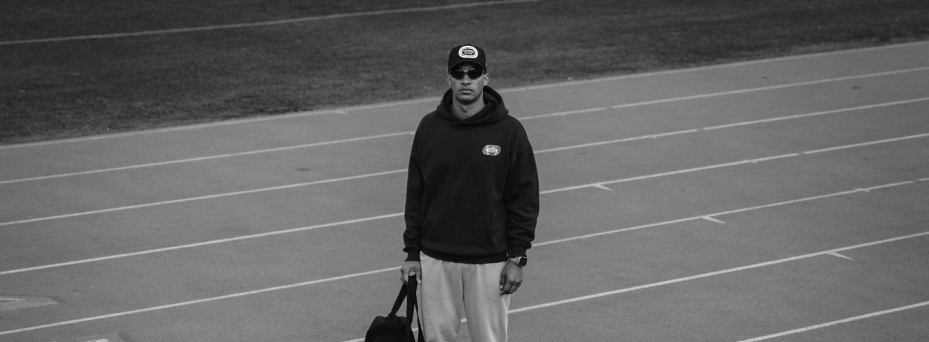 Person standing on a track holding a bag, wearing a huega house hat and hoodie